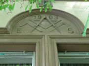 Détail de la façade qui se trouve au rez-de chaussée à gauche au dessus des fenêtres sur rue avec les initiales de l'architecte, des outils sculptés (équerre et compas) et la date de construction 1903.